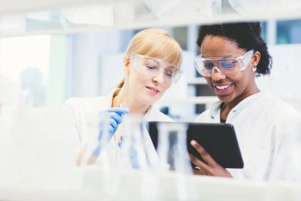 Two female scientists in safety goggles examining a blue liquid sample and tablet in a laboratory